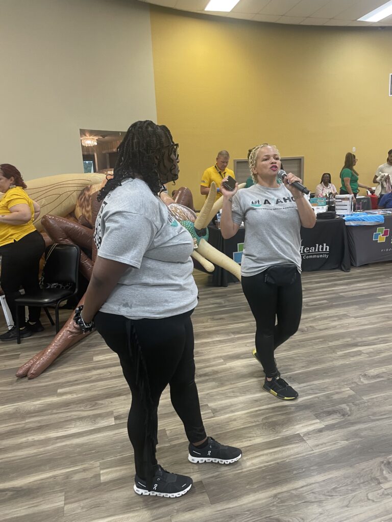 Two women from the African American Health Coalition, including Board Chair Jeanette Lastrape, stand at the front of the room during the Wellness on Wheels event. One holds a microphone and addresses attendees, welcoming them to the event. Both wear matching AFAHC t-shirts and black sneakers.